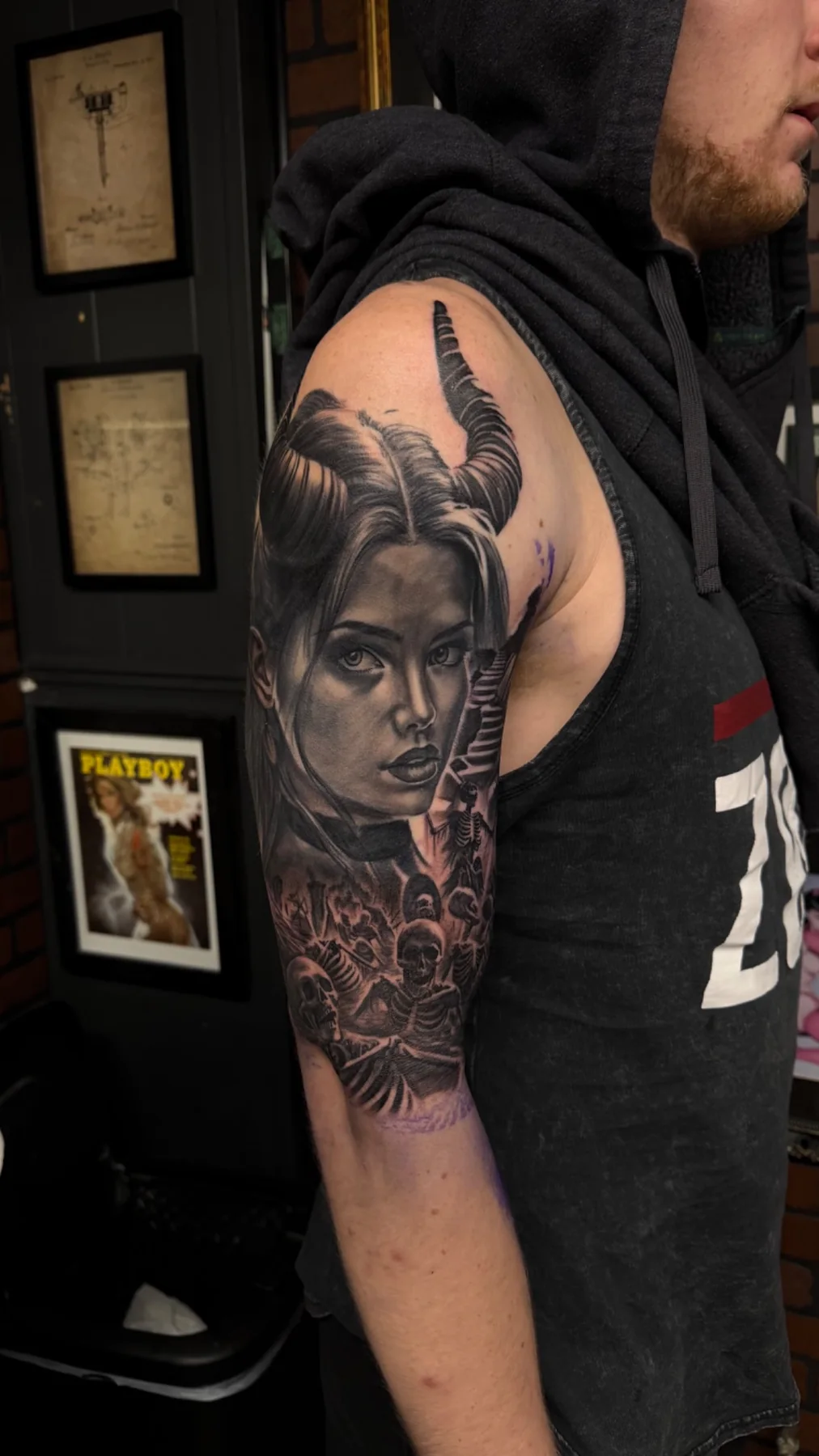 Black and grey realism upper-arm tattoo featuring a horned female portrait and skeletal scene